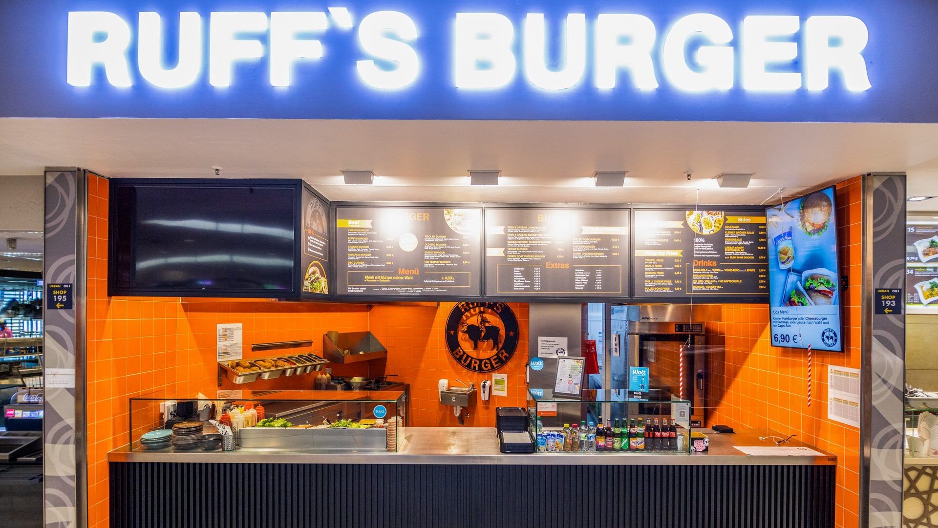 Innenansicht von "Ruff's Burger" mit Theke, Küche im Hintergrund, orangener Wand, beleuchtetem Logo und Menütafeln.