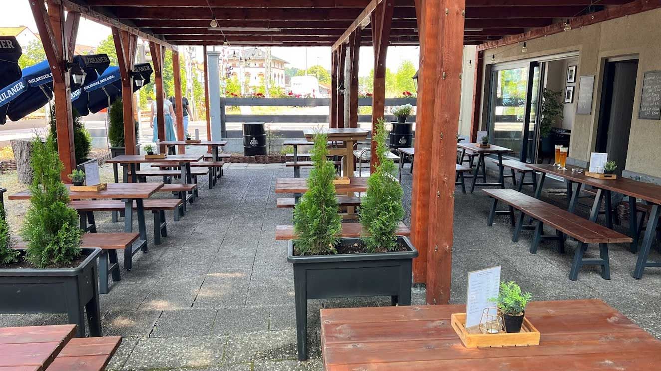 Überdachte Außenterrasse von "Burger House" mit Holzbänken, Tischen und in Reihen platzierten Pflanzkübeln.