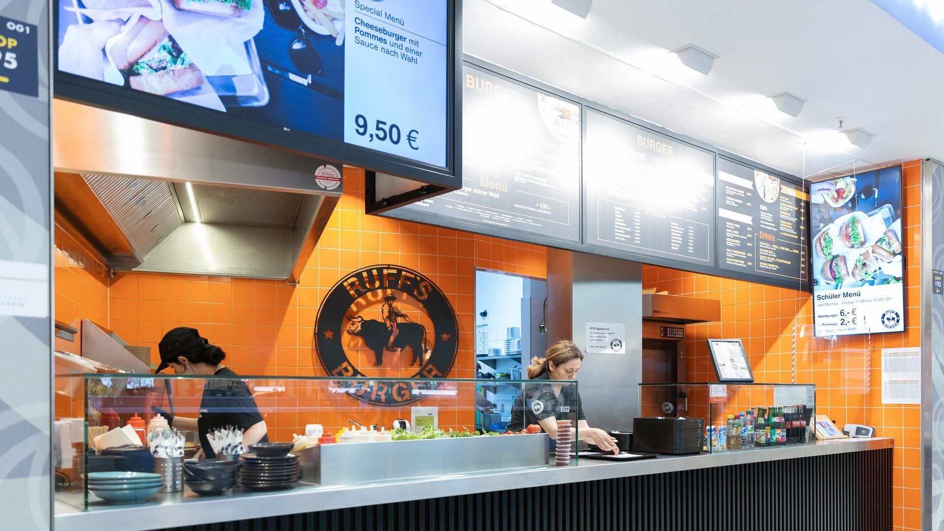 Innenansicht von "Ruff's Burger" mit Theke, Mitarbeiter bei der Zubereitung, Küche, orangener Wand und Menütafeln.
