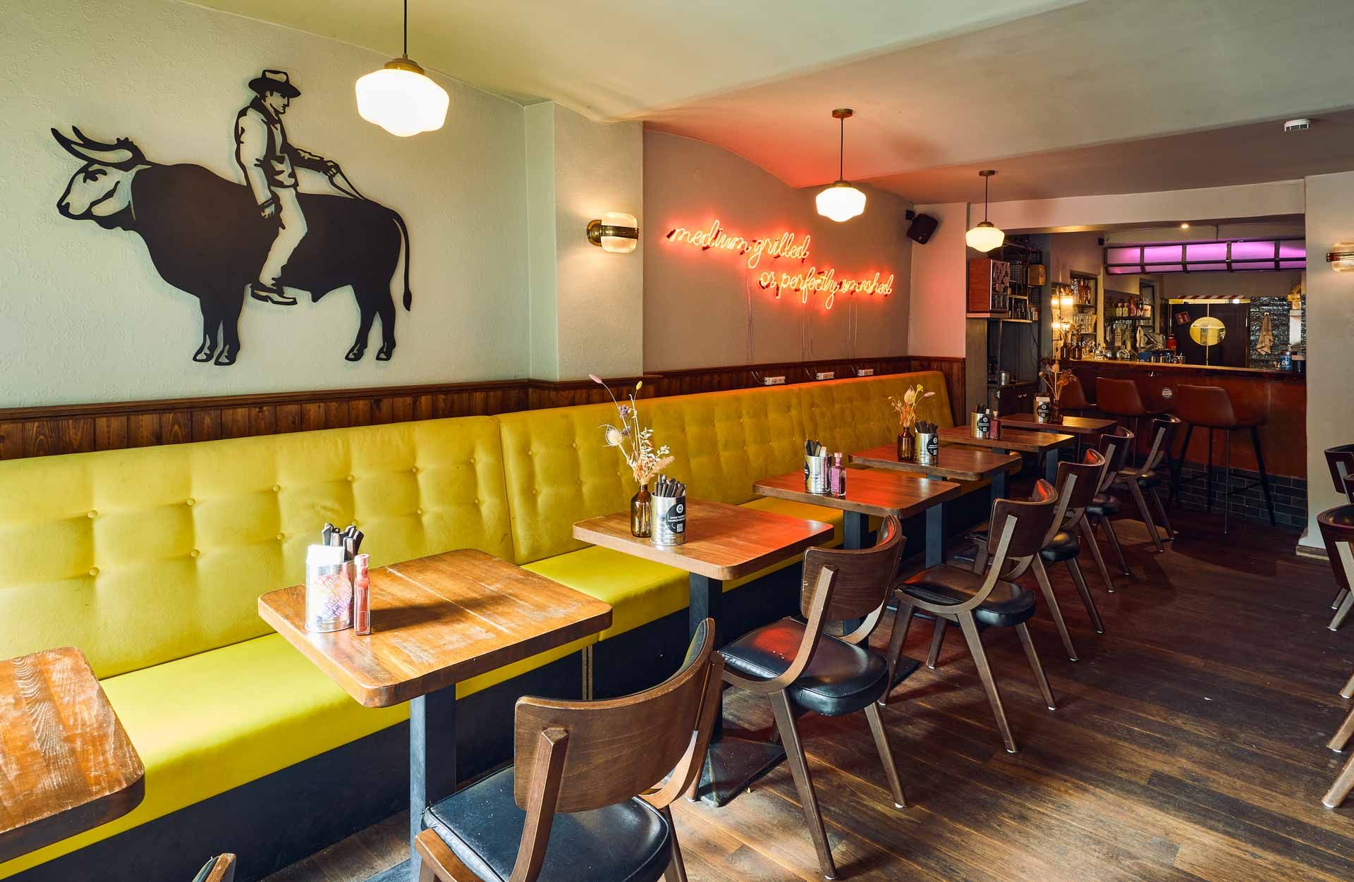Eingangsbereich von "Otto's Burger" mit gelber Polsterbank, Holztischen, Holzstühlen, Dekoration und Blick Richtung Barbereich.