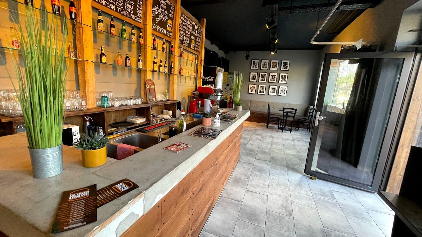 Innenraum von Burger House mit Theke aus Holz und Beton, Flaschenregalen, Speisetafeln und Sitzbereich im Hintergrund.