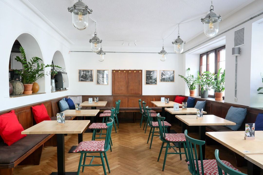 Innenansicht von "Ruff's Burger" mit Holztischen, grünen Stühlen, Parkettboden, weißen Wänden, Wandbildern und Pflanzen auf den Fensterbänken.