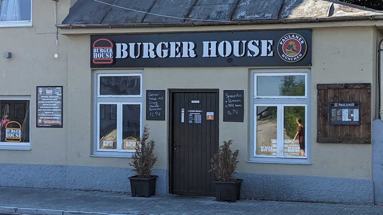 Außenansicht von "Burger House" mit zentraler Eingangstür, Fenstern, Schildern und Logo über dem Eingang.