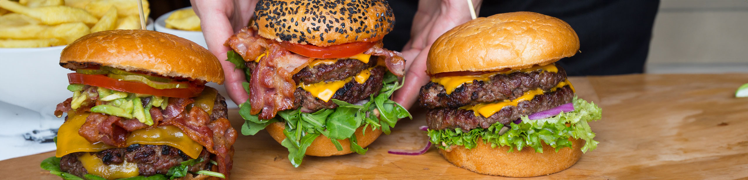 Drei große Burger, die auf einem Holzbrett angerichtet wurden. Der mittlere Burger wird von zwei Händen hochgehoben. Die Burger sind alle mit vielen Zutaten belegt. Im Hintergrund steht eine Schale Pommes.