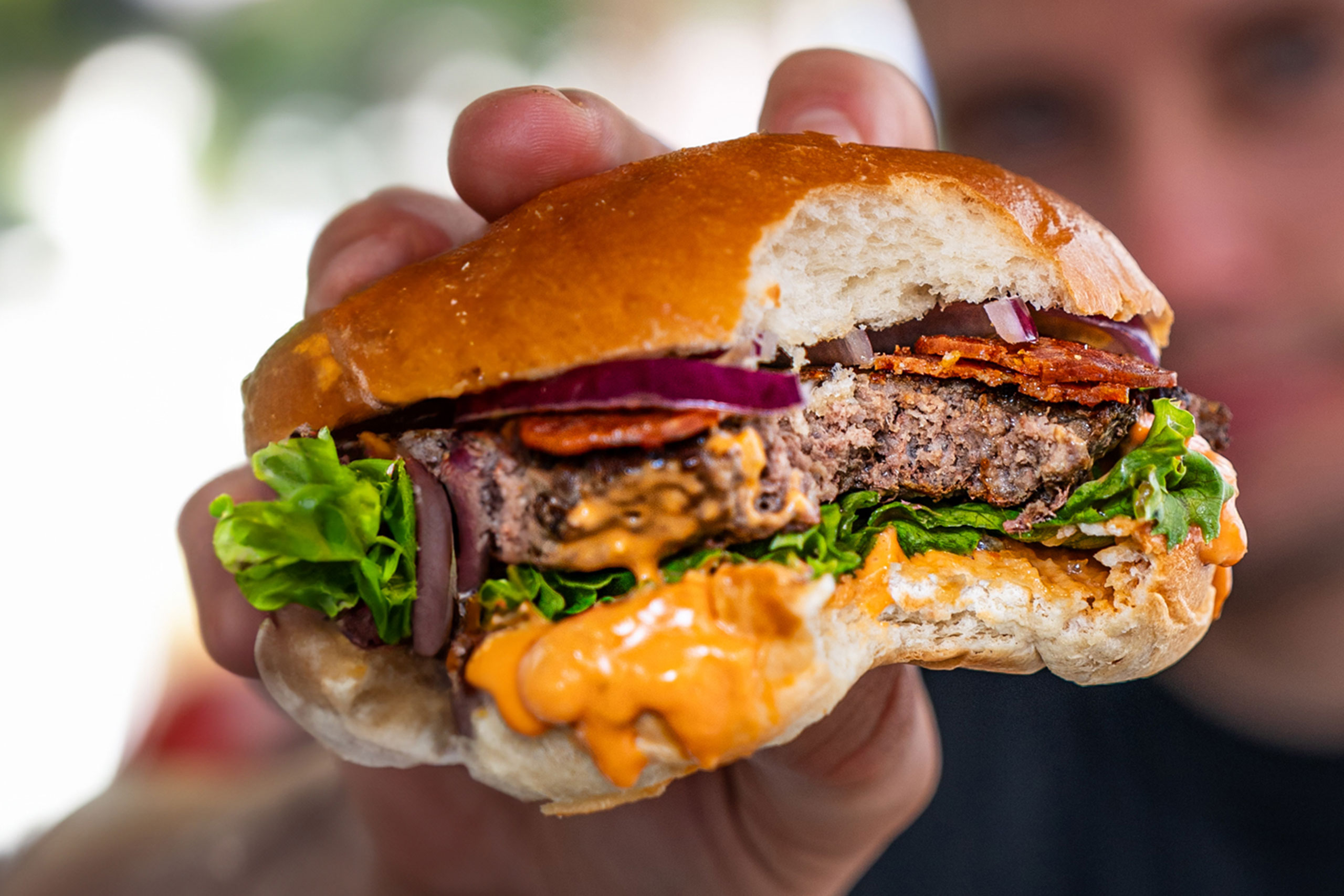 Nahaufnahme eines angebissenen Burgers mit Salat, Zwiebeln, Bacon, Patty und Sauce im Brioche-Bun.