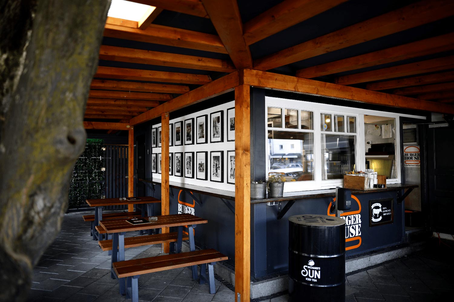 Überdachter Sitzbereich des „Burger House“ mit Holzbänken, dekorativer Fotowand und Blick auf den Bestellcounter mit geöffnetem Fenster.