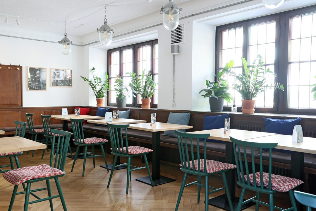 Innenansicht von "Ruff's Burger" mit Holztischen, Fensterfront, Pflanzen auf der Fensterbank, grünen Stühlen und roten Kissen.