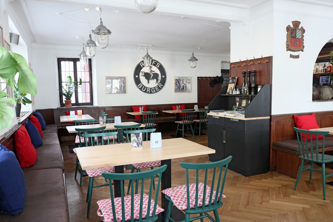 Innenansicht von "Ruff's Burger" mit Holztischen, grünen Stühlen mit roten Polsterkissen, Wandlogo und Parkettboden. Am Rand eine Servicestation mit Menükarten und Salz-, und Pfefferstreuern.