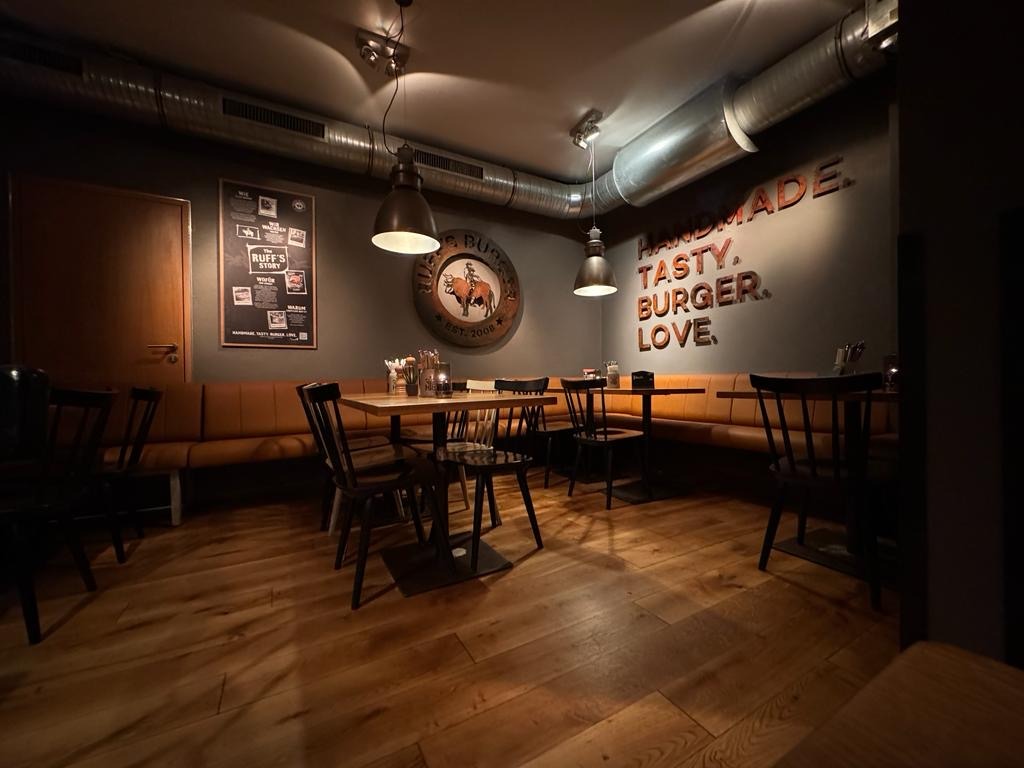 Innenansicht von "Ruff's Burger" mit Holzboden, dunklen Wänden, brauner Polsterbank, Wandlogo und Schriftzug "Handmade Tasty Burger Love".