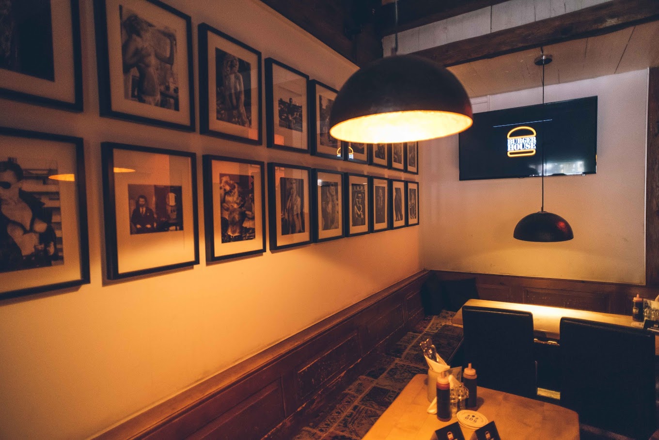Innenbereich des Restaurants mit Holztisch, Wandgalerie aus Schwarz-Weiß-Fotos und TV-Bildschirm mit Logo.