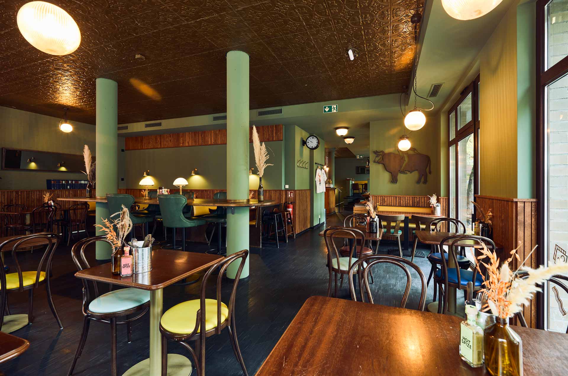 Innenansicht von "Otto's Burger" mit Holztischen, Stühlen, großen Fenstern, blauen Säulen und gemusterte Decke.